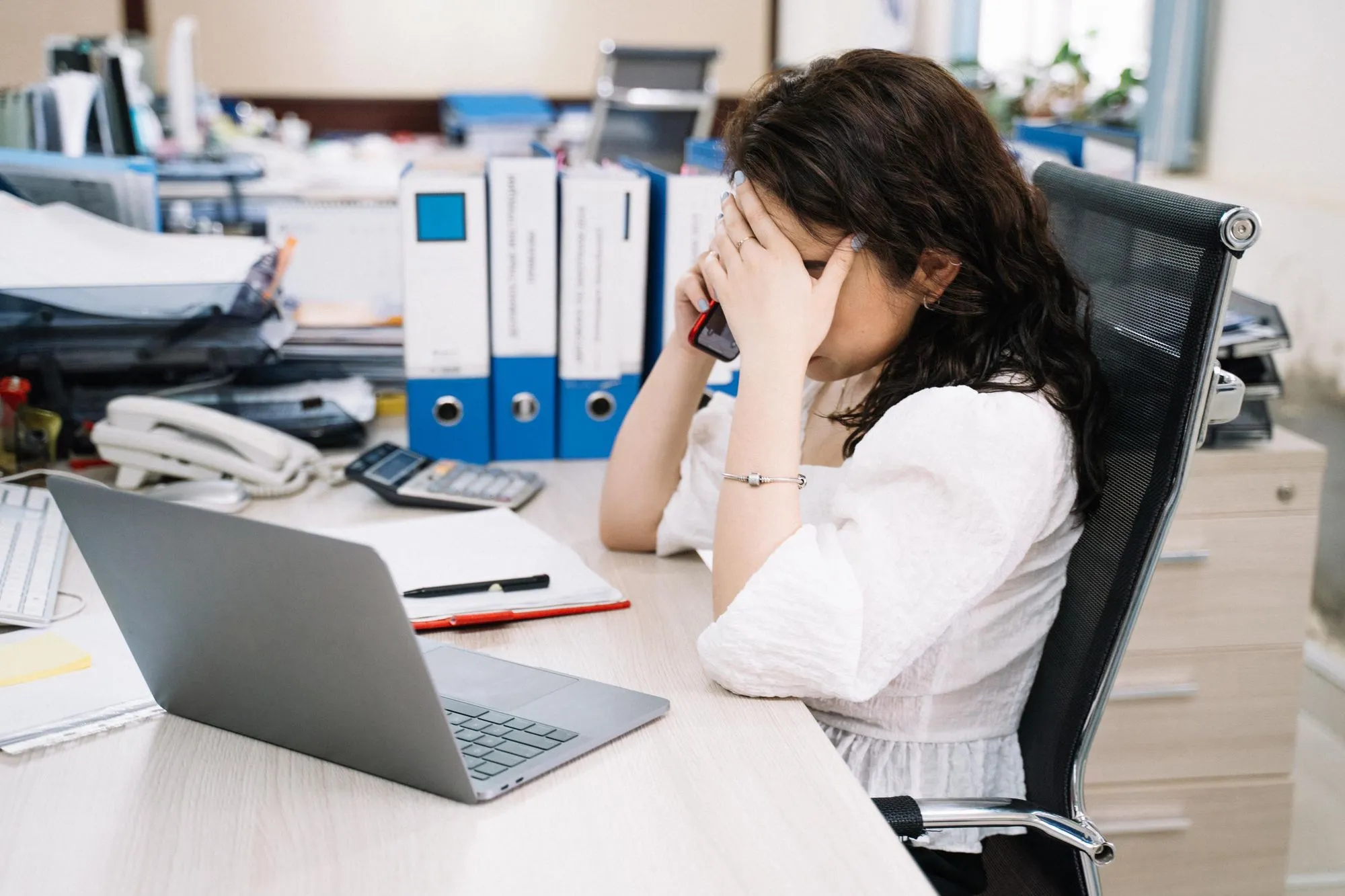 An office worker at a cluttered desk with a phone to her ear, binders and paperwork stacked around her